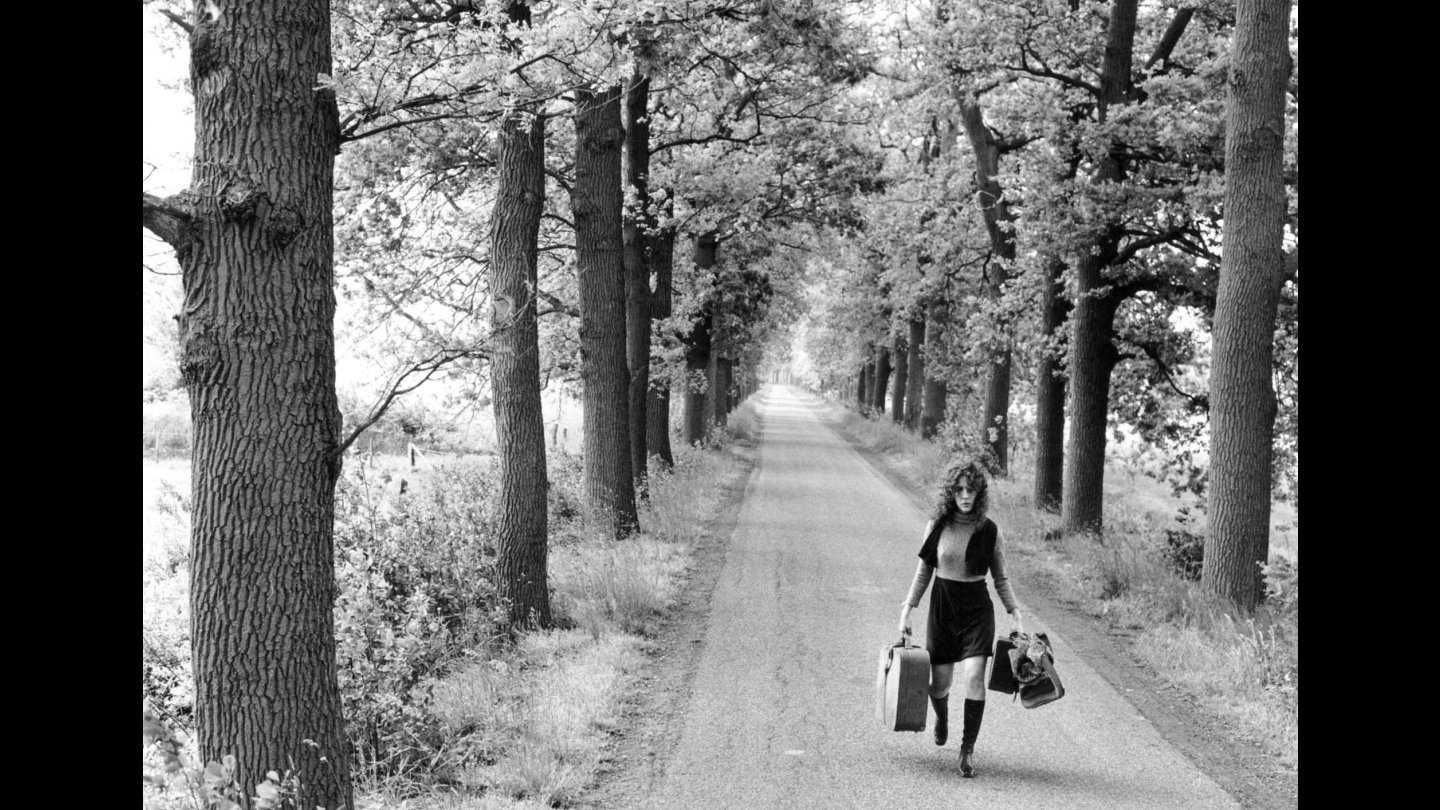 Filmstill von "Boven de Bergen", Frau in schwarzem Kleid und Stiefeln geht auf einer von Bäumen gesäumten Landstraße und trägt zwei Koffer