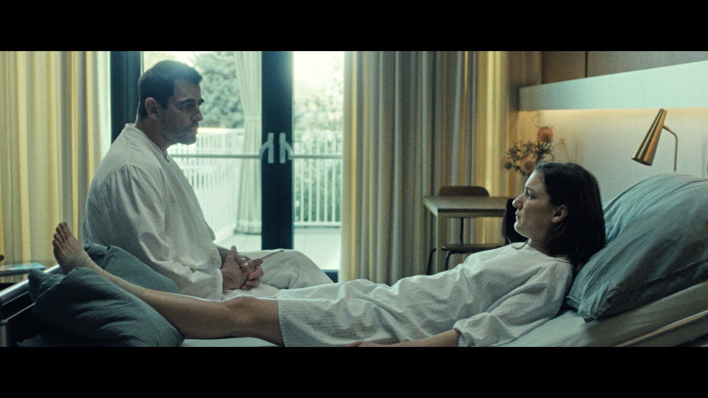 Filmstill aus MOTHER'S BABY: Eine Frau liegt auf einem Krankenhausbett und blickt zu einem Mann, der auf dem Bettende sitzt.
