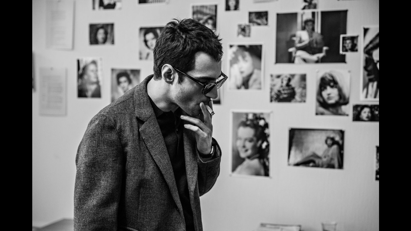 Filmstill aus NOUVELLE VAGUE: Das Bild ist schwarz-weiß. Ein Mann mit Brille zieht an einer Zigarette, er trägt Sonnenbrille und ist zur Seite gerichtet. Im Hintergrund, an der Wand, befinden sich viele Fotos und Bilder.