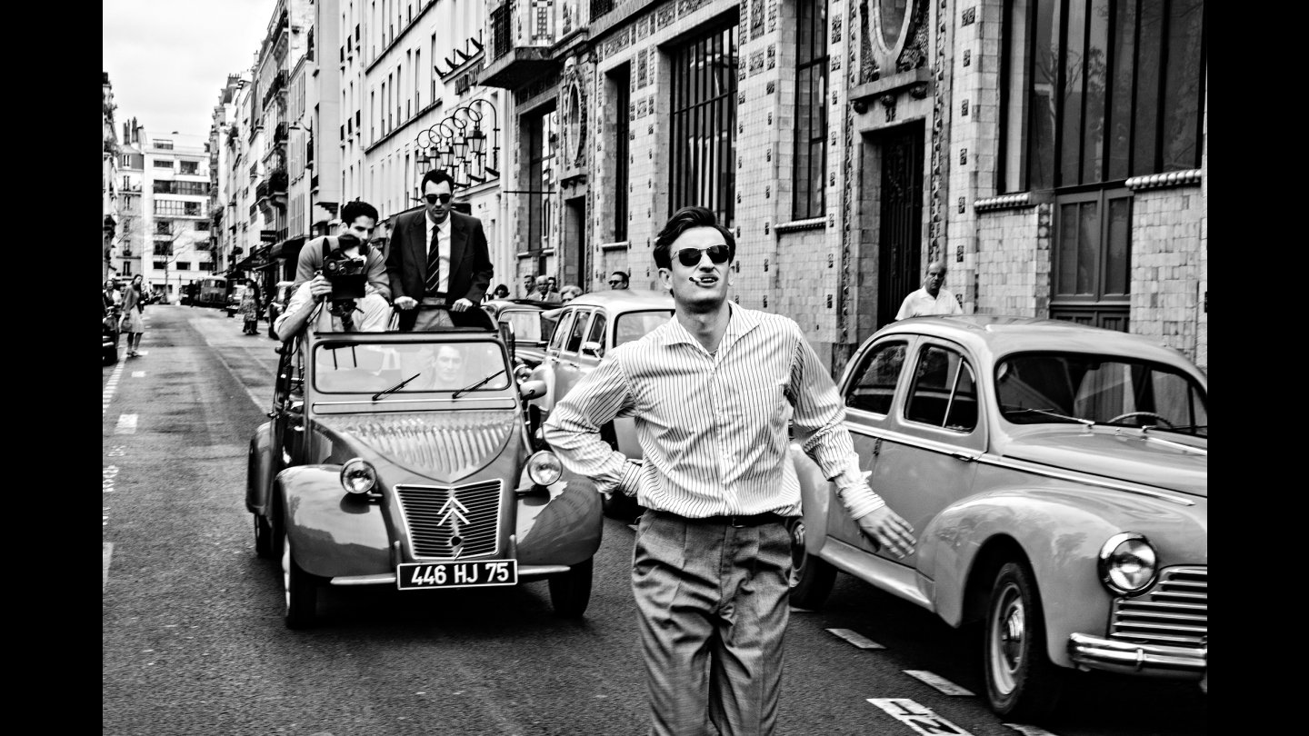 In Schwarz Weiß: In einer Gasse mit parkenden Autos geht ein Mann mit Sonnenbrille und Zigarette im Mund die Straße entlang. Hinter ihm fährt eine Gruppe Männer mit einem Oldtimer Auto und fotografieren ihn von hinten.