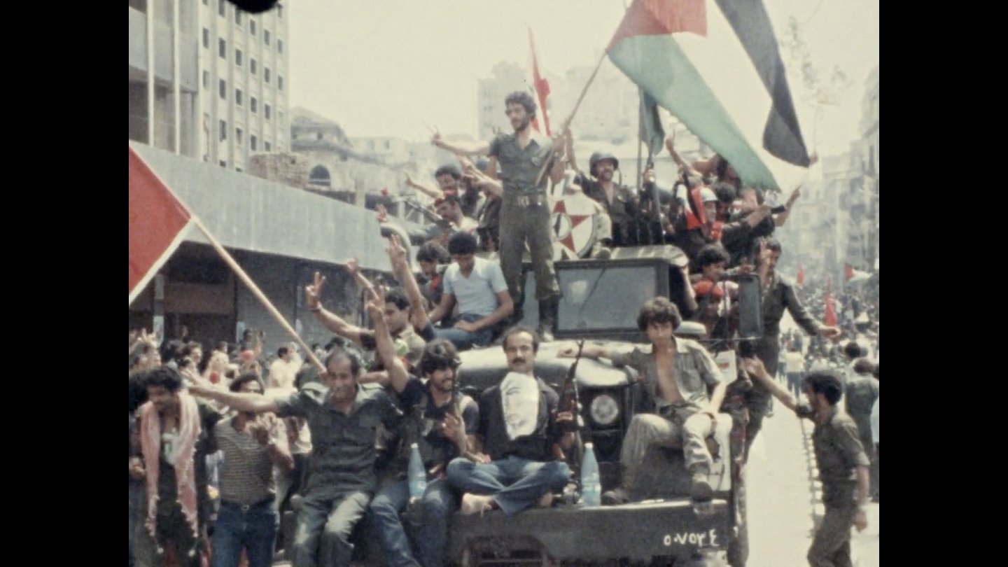 FIlmstill Jocelyne Saab: Gruppe von Menschen auf einem Fahrzeug in einer Stadt, einige halten palästinensische Fahnen und zeigen Friedenszeichen