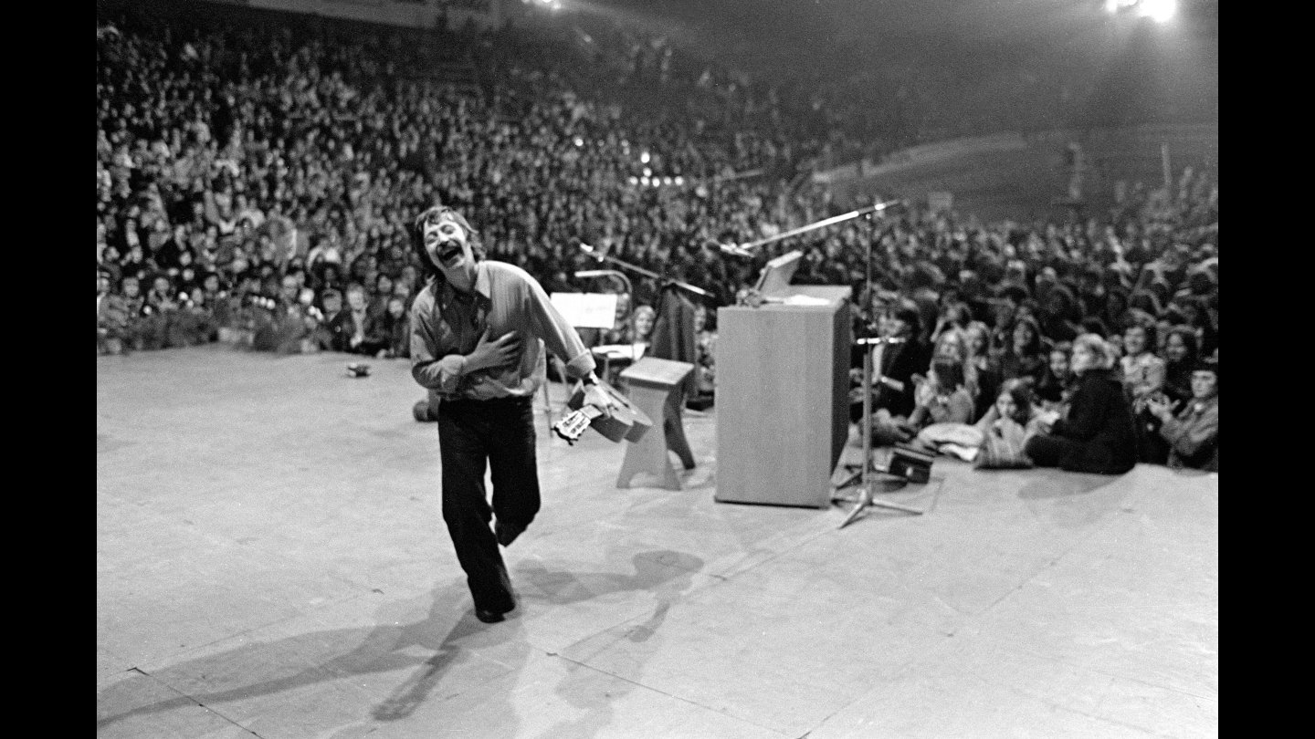 Wolf Biermann mit Mikrofon und Zetteln in der Hand läuft lachend auf einer Bühne vor großem Publikum. Wolf Biermann bei seinem berühmtesten Konzert: In der Kölner Sporthalle im November 1976. Drei Tage spaeter wurde er aus der DDR ausgebuergert.