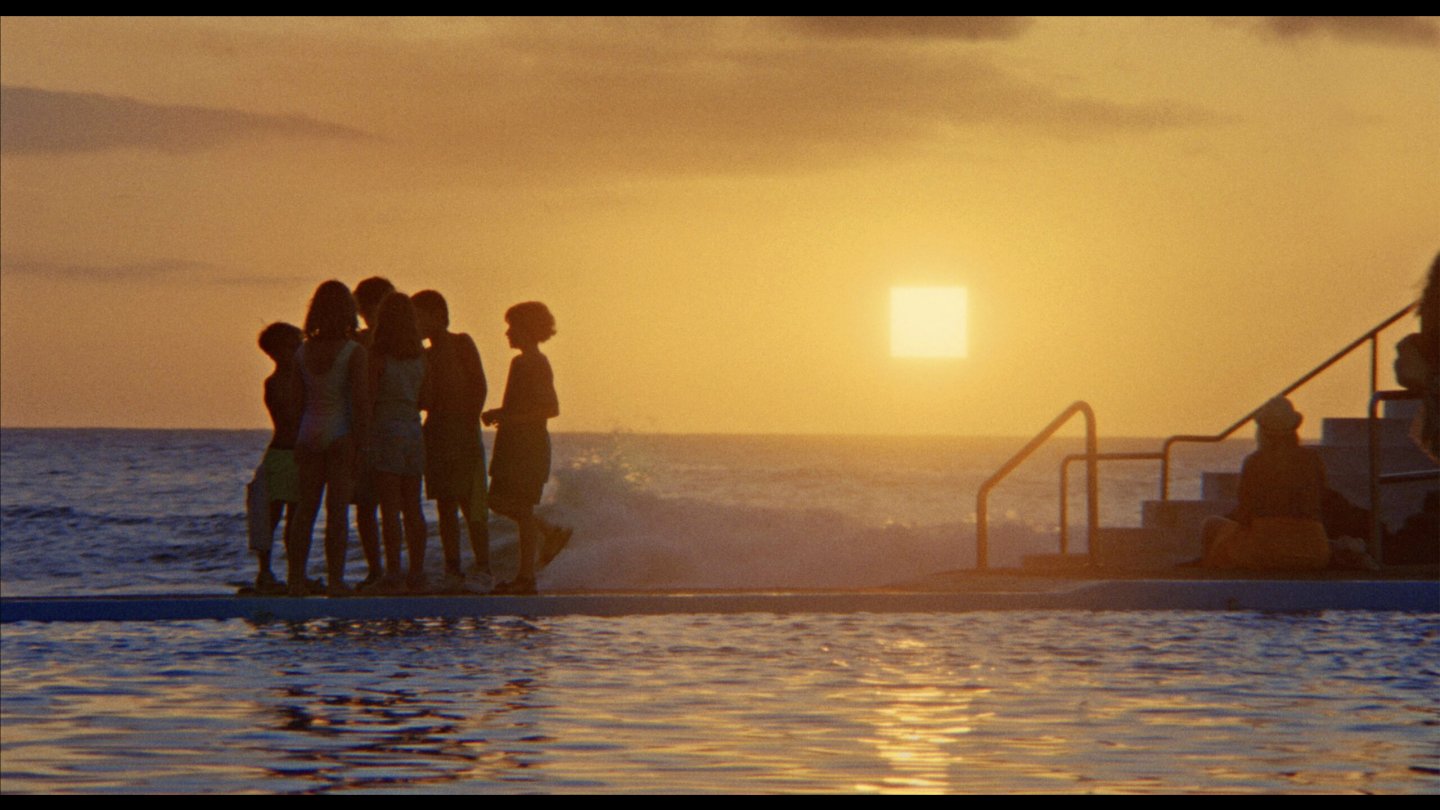 Gruppe von Menschen steht am Rand eines Pools mit Blick auf das Meer bei Sonnenuntergang