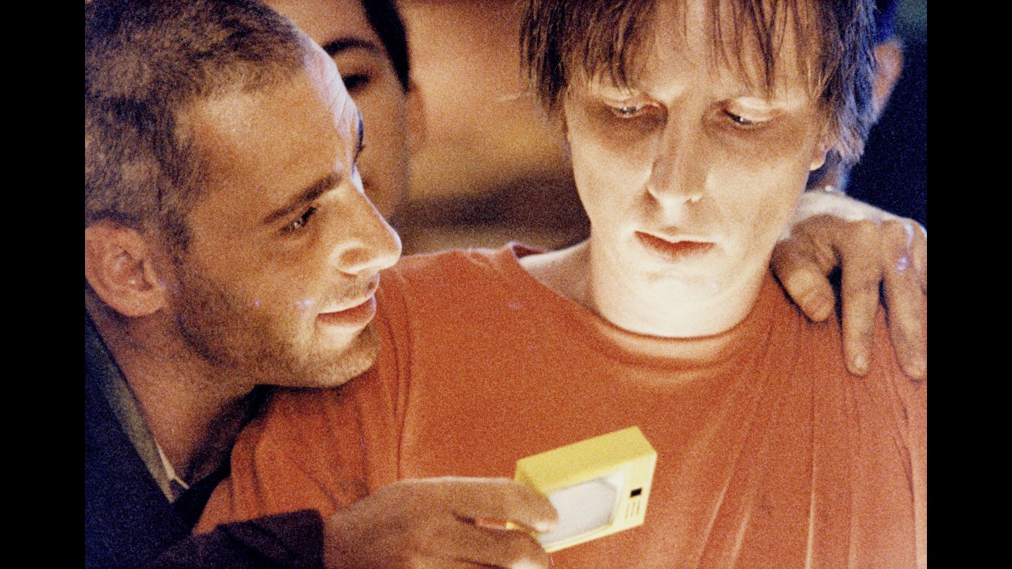 Filmstill aus HALBE WELT. Mann mit kurz geschorenem Haar trägt runde Sonnenbrille mit hellen Bügeln und brauner Jacke