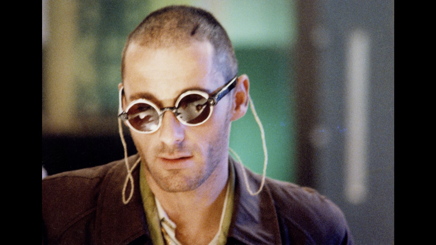 Filmstill HALBE WELT. Mann mit kurz geschorenem Haar trägt runde Sonnenbrille mit hellen Bügeln und brauner Jacke