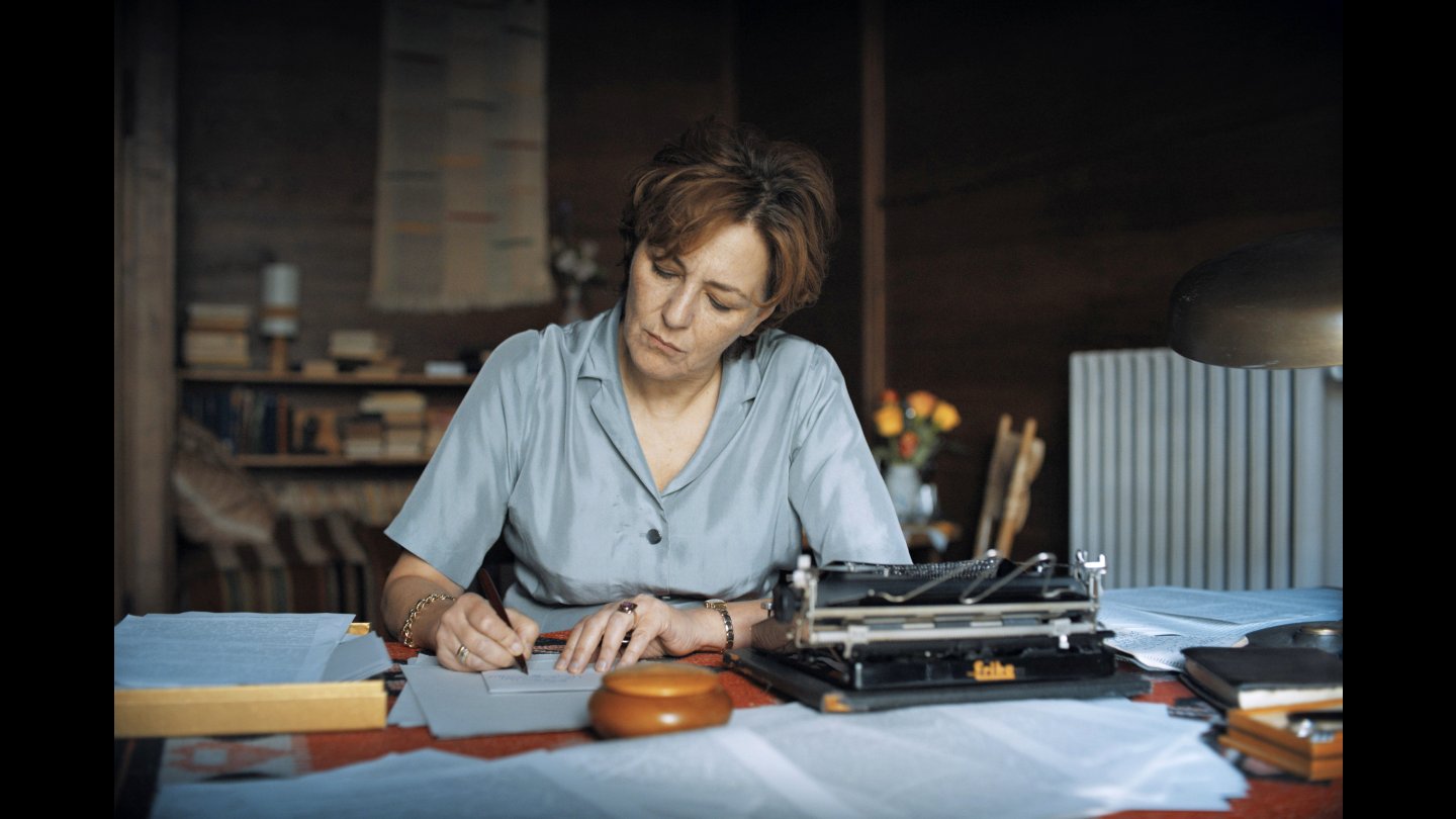 Susanne Wolff (die Frau des Architekten) sitzt an einem Tisch und schreibt auf Papier neben einer Schreibmaschine in einem Raum mit Bücherregal und Lampe