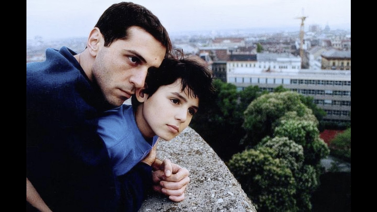 FIlmstill JUGO FILM. Zwei Personen lehnen an einer Mauer und blicken nachdenklich in die Ferne über eine Stadt mit Bäumen und Gebäuden