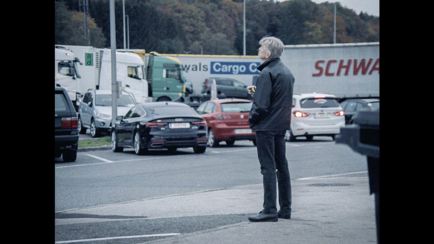 Ein älterer Mann mit grauen Haaren steht auf einem Parkplatz und blickt nach oben, im Hintergrund stehen mehrere Lastwagen und Autos.