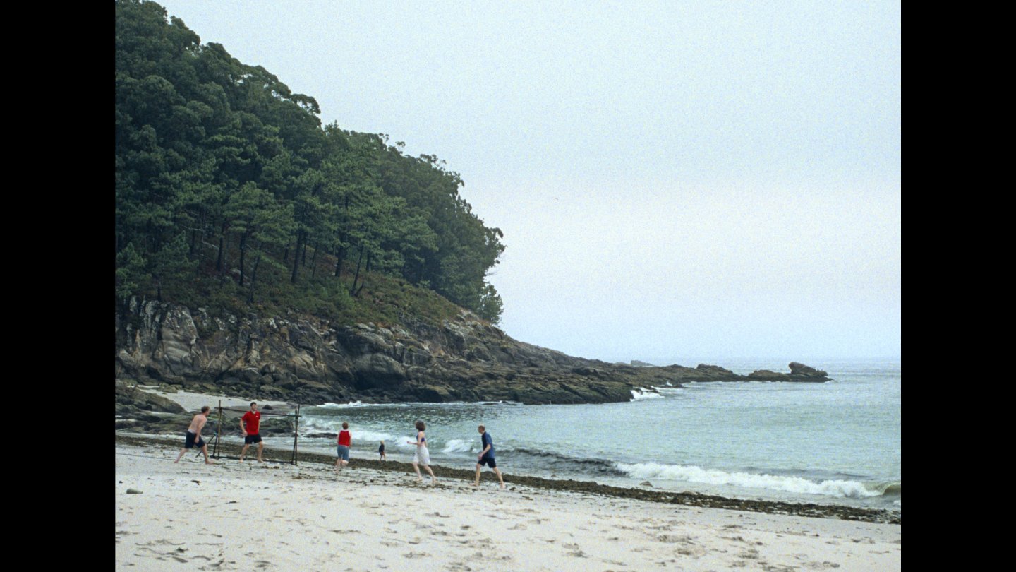 Mehrere Personen spielen am Strand vor bewaldeten Klippen Fußball