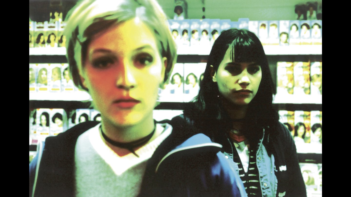 Filmstill SLIDIN' – ALLES BUNT UND WUNDERBAR. Zwei junge Frauen stehen vor einer Wand mit vielen kleinen Fotos, die vordere Frau trägt ein helles Oberteil und einen schwarzen Choker, die hintere Frau hat dunkle Haare und trägt eine Jacke über einem gestreiften Shirt