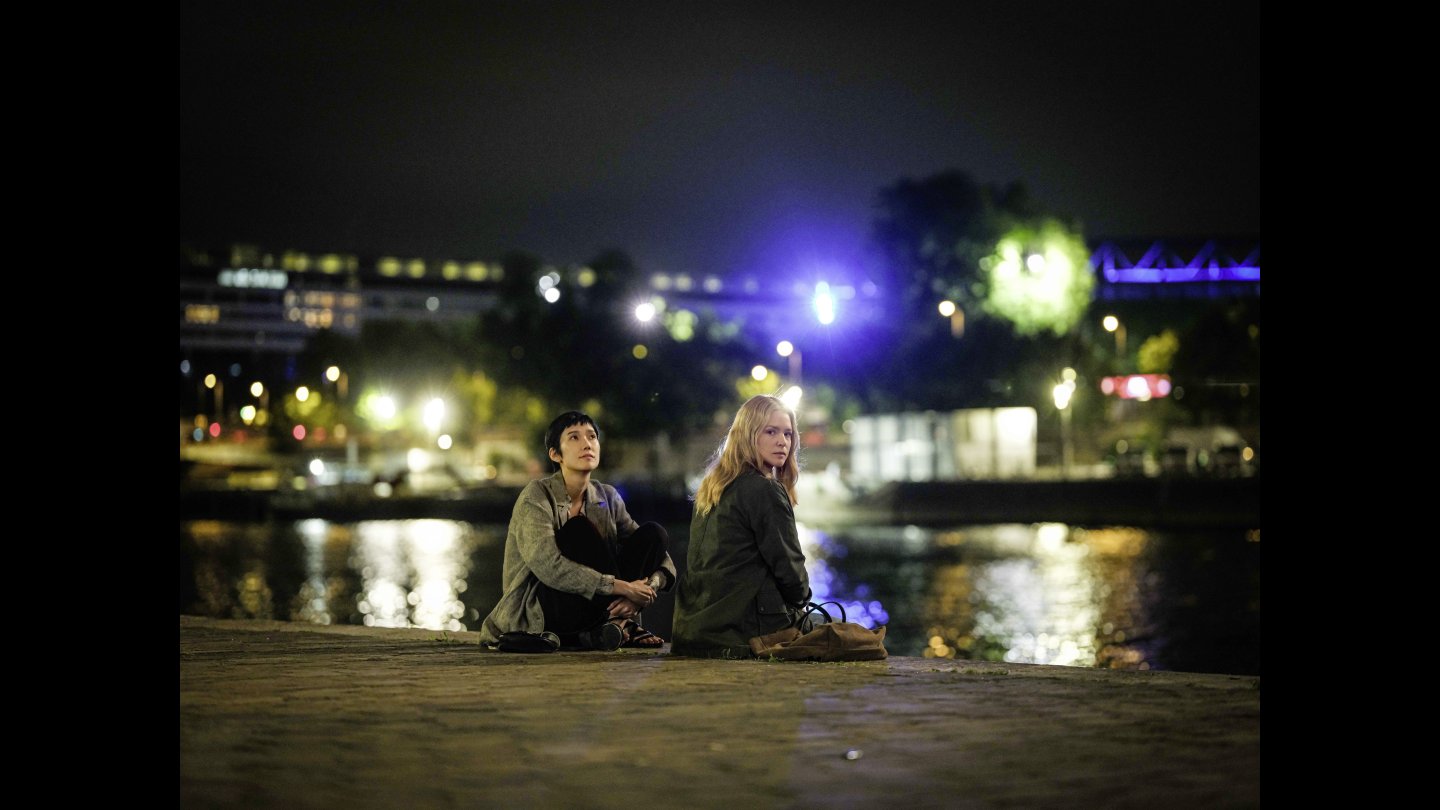 Zwei Frauen sitzen am Boden vor einem Fluss, um sie herum die Lichter der Stadt. Eine der Frauen blickt in die Kamera.