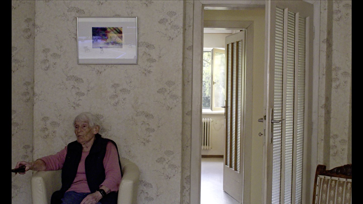 Eine ältere Frau sitzt auf einem gemütlichen Stuhl, in der Hand hat sie eine Fernbedienung. Sie befindet sich in einer Wohnung und einem Haus.
