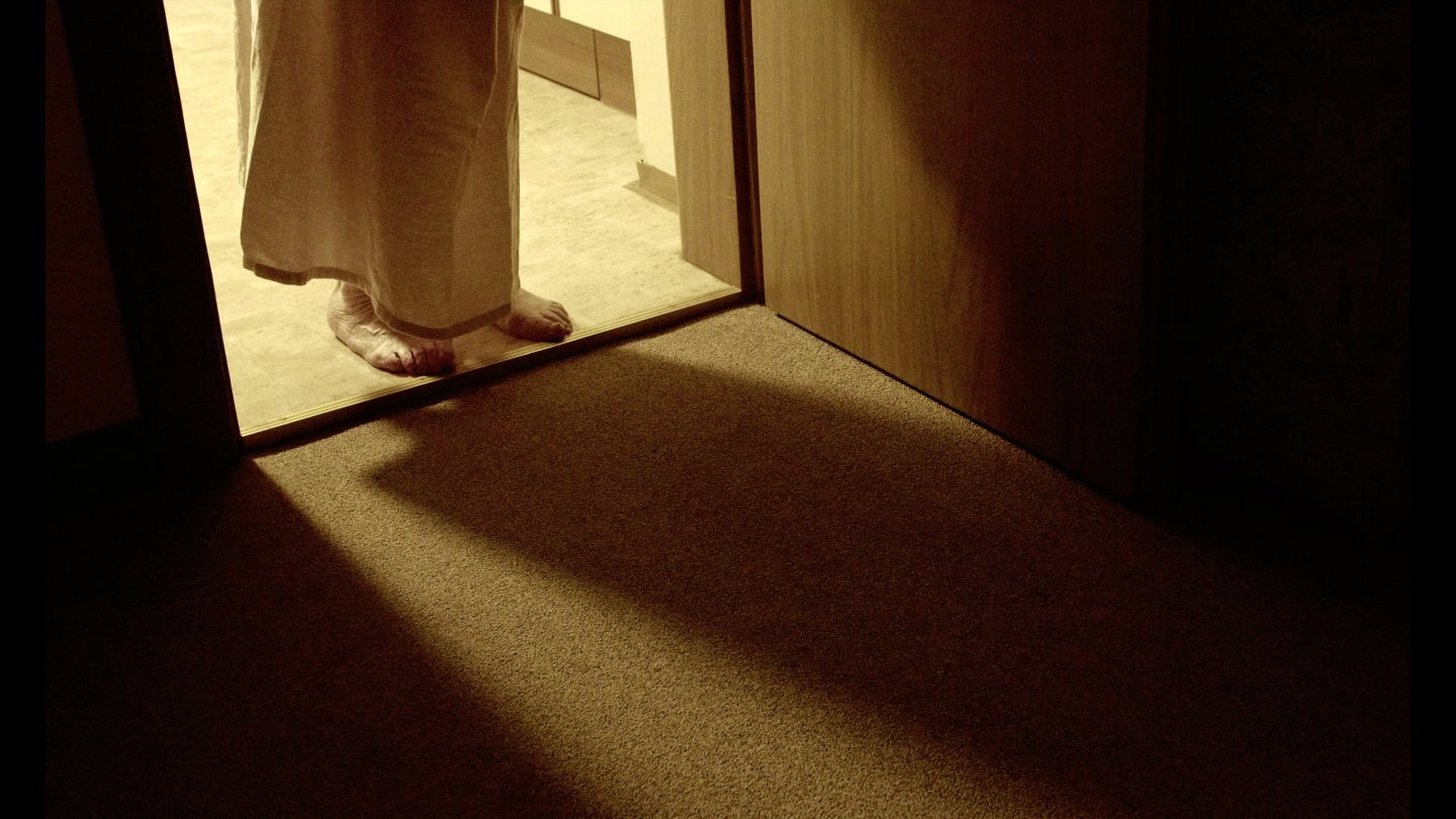 Barfüßige Person steht in einem hell erleuchteten Türrahmen mit langem Kleid und wirft Schatten auf einen dunklen Teppichboden
