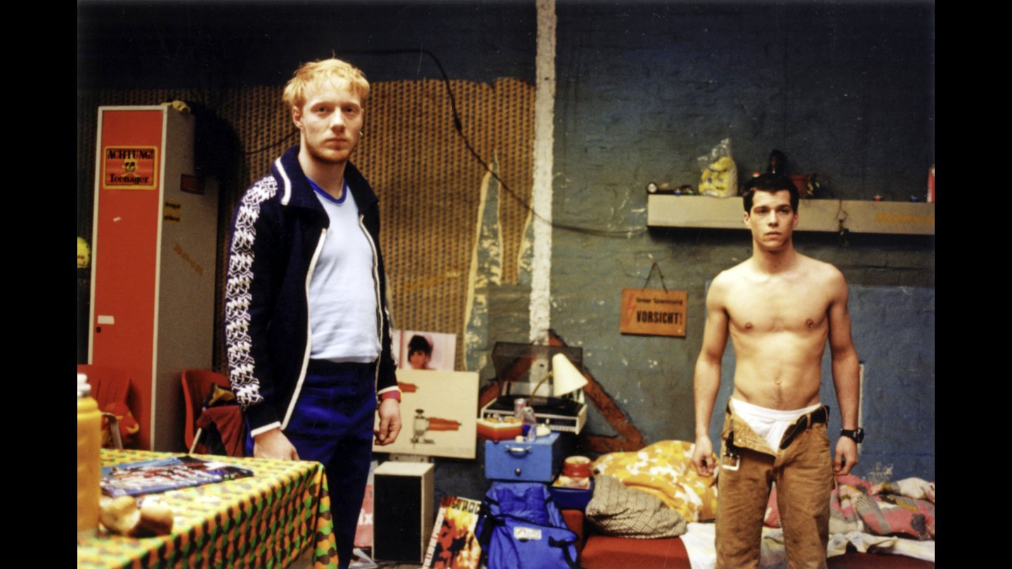 Filmstill TEMPO. Zwei junge Männer stehen in einem unordentlichen Raum mit buntem Tisch, Kisten und Warnschildern an der Wand, einer trägt Trainingsjacke und Hose, der andere ist oberkörperfrei mit oben offener brauner Hose. Man sieht die Unterhose