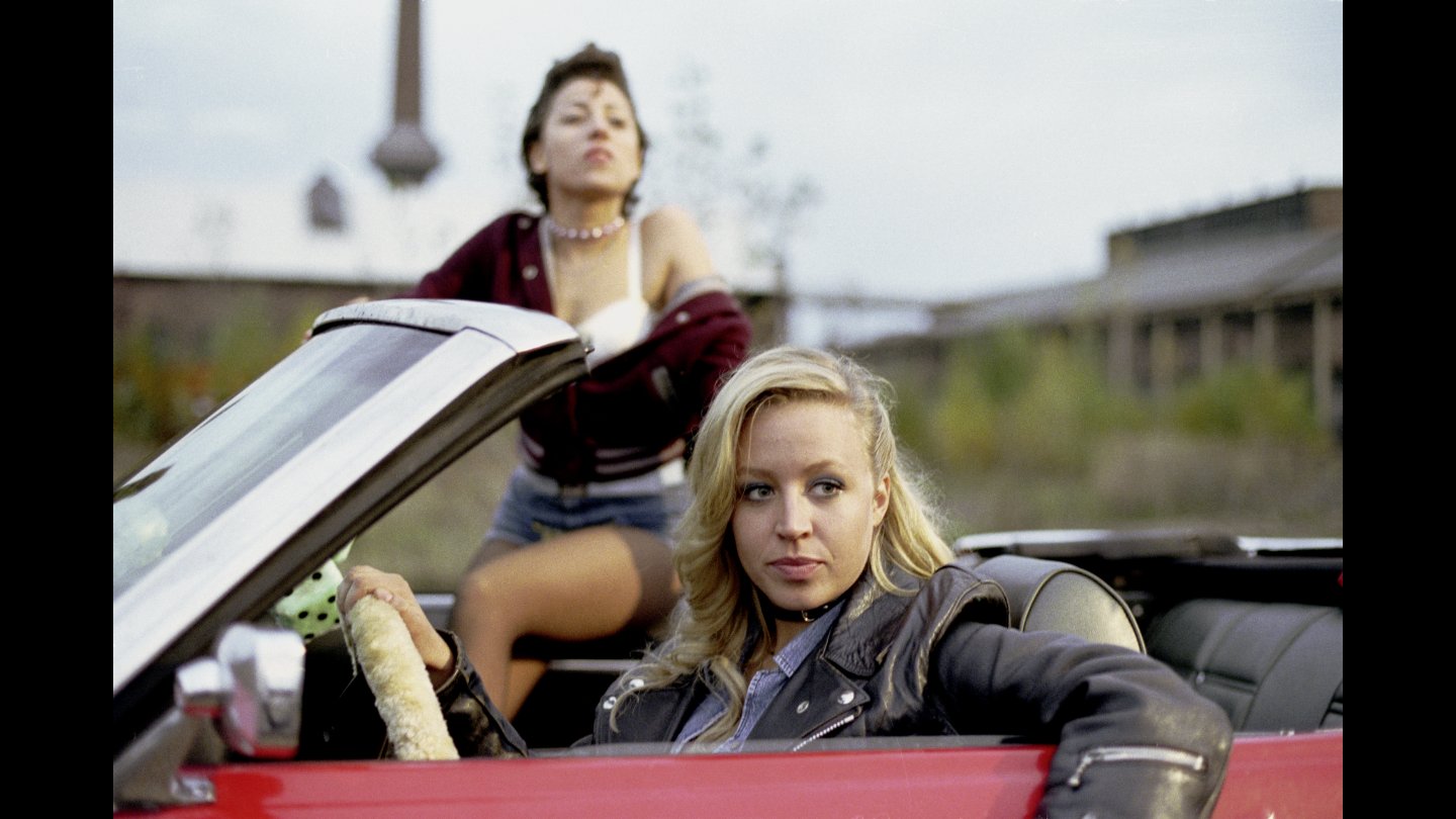 Filmstill TERNITZ, TENNESSEE Zwei Frauen in einem roten Cabrio, eine sitzt am Steuer und schaut nach rechts, die andere steht hinter dem Auto