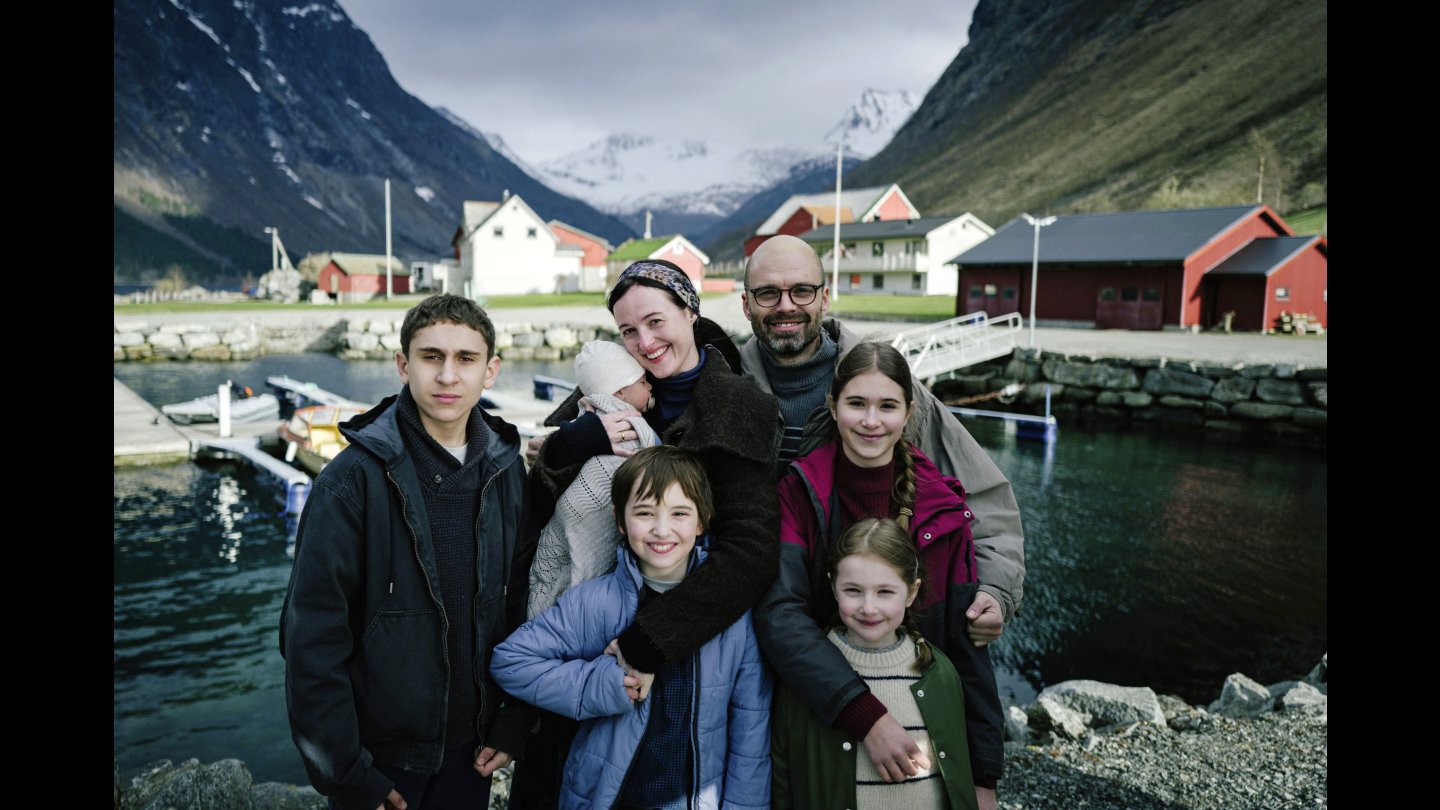 Ein Familienporträt, abgebildet sind fünf Kinder und 2 Erwachsene. Sie stehen vor einem Gewässer und lächeln in die Kamera.
