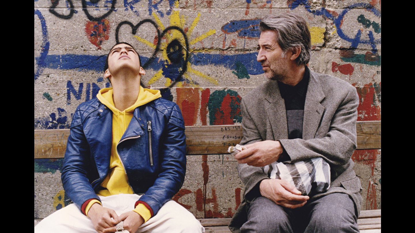 Filmstill JUGOFILM: Zwei Menschen sitzen nebeneinander, hinter ihnen eine mit Graffiti bemalte Wand.
