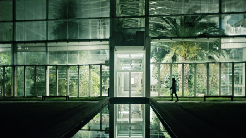 Filmstill "Bestiari, Erbari, Lapidari", Person geht in einem modernen, verglasten Gebäude mit Blick auf Palmen und Pflanzen im Inneren
