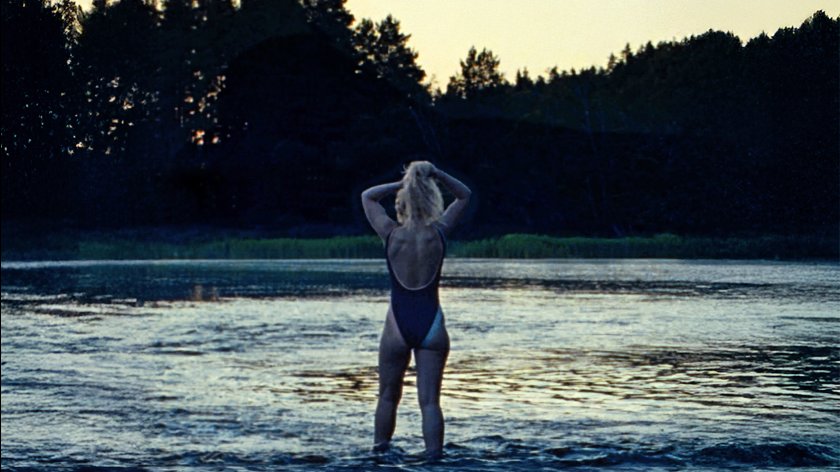 Eine Frau steht in einem See, mit dem Rücken zur Kamera. Sie fasst sich mit beiden Händen in die Haare.