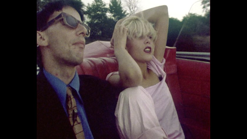Filmstill von "Diorissimo"; Frau in rosa Kleid sitzt entspannt im Auto, hebt Arme hinter den Kopf, Mann in Anzug und Krawatte sitzt daneben