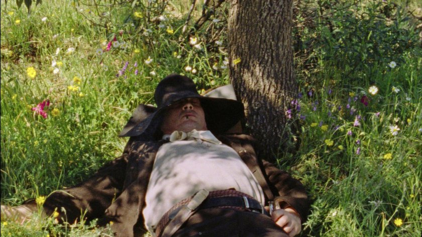 Filmstill aus AS ESTAÇÕES: Ein Mann unter einem Baum im Gras, er hat einen Hut über den Kopf liegen. Die Sonne scheint, der Mann liegt im Schatten.