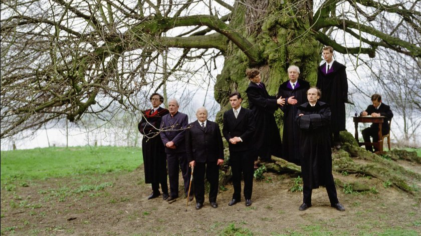 Filmstill aus "Zechmeister"; Gruppe von neun Personen in schwarzen Anzügen und Roben steht vor einem großen, kahlen Baum auf einer Wiese, eine Person sitzt an einem Holztisch rechts