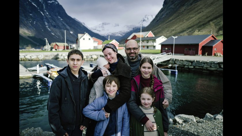 Ein Familienporträt, abgebildet sind fünf Kinder und 2 Erwachsene. Sie stehen vor einem Gewässer und lächeln in die Kamera.