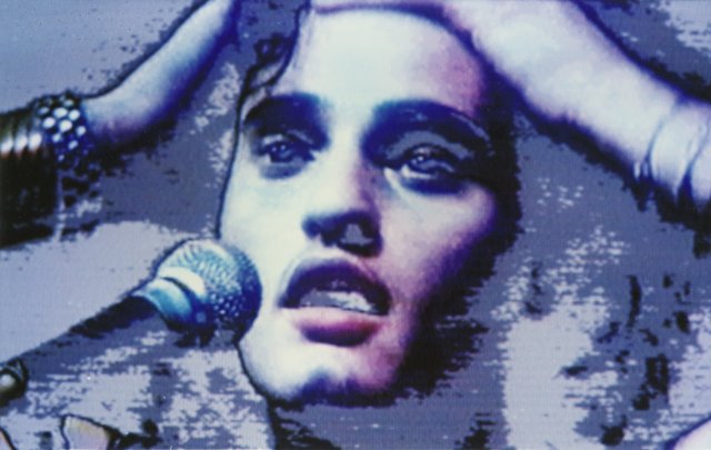 Filmstill aus ROCKTÉRÍTŐ: Nahaufnahme von einer Person, die in ein Mikrofon singt, die Hände an den Kopf gelegt.