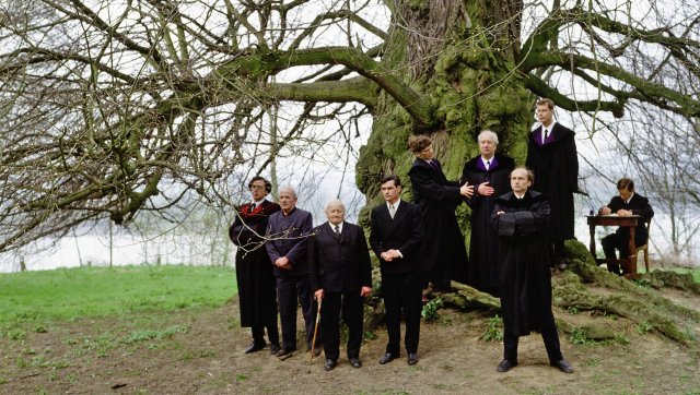 Filmstill aus "Zechmeister"; Gruppe von neun Personen in schwarzen Anzügen und Roben steht vor einem großen, kahlen Baum auf einer Wiese, eine Person sitzt an einem Holztisch rechts