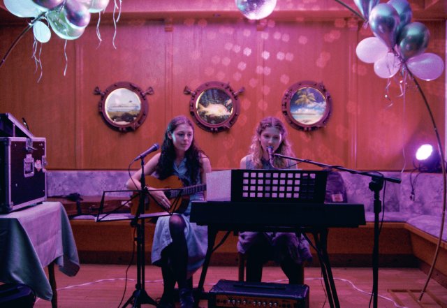 Zwei Frauen sitzen auf einer Bühne, eine spielt Gitarre, die andere Klavier und singt, umgeben von Luftballons und maritim dekoriertem Hintergrund mit Bullaugen.
