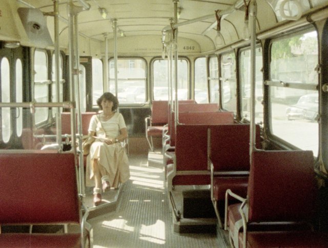 Eine Frau sitzt in einem Bus oder einer Straßenbahn, alle Plätze um sie herum sind leer. Die Sitze sind rot.