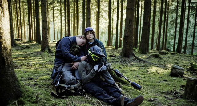 Drei Personen in Outdoor-Kleidung sitzen im Wald auf moosbedecktem Boden, eine Person lehnt den Kopf an die Schulter einer anderen