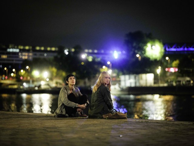Zwei Frauen sitzen am Boden vor einem Fluss, um sie herum die Lichter der Stadt. Eine der Frauen blickt in die Kamera.
