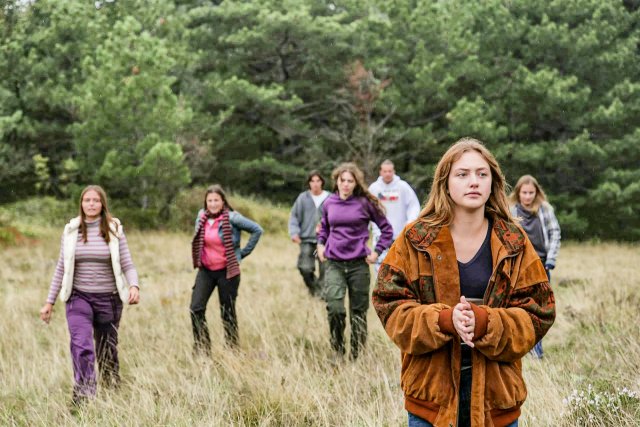 Eine Gruppe von Menschen wandert durch ein Feld, im Vordergrund steht eine junge Frau. Es wirkt herbstlich.