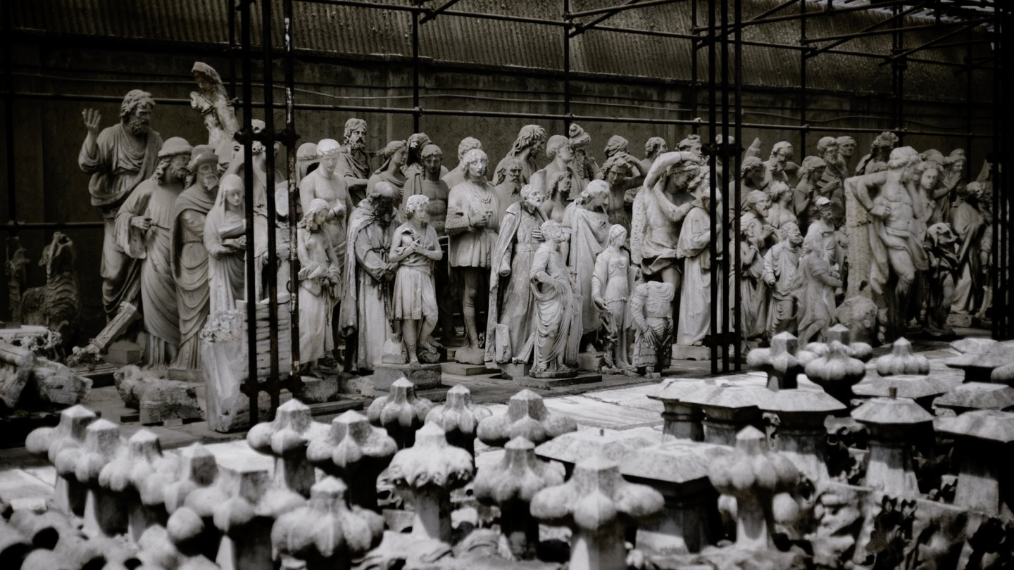 Filmstill aus L’INFINITA FABBRICA DEL DUOMO. Abgebildet sind etliche Statuten, alle knapp nebeneinander in einem Raum stehen.