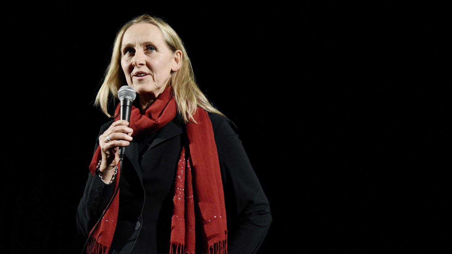 Angela Summereder mit Schulterlangem blonden Haar, schwarzem Mantel und rotem Schal hält ein Mikrofon vor schwarzem Hintergrund