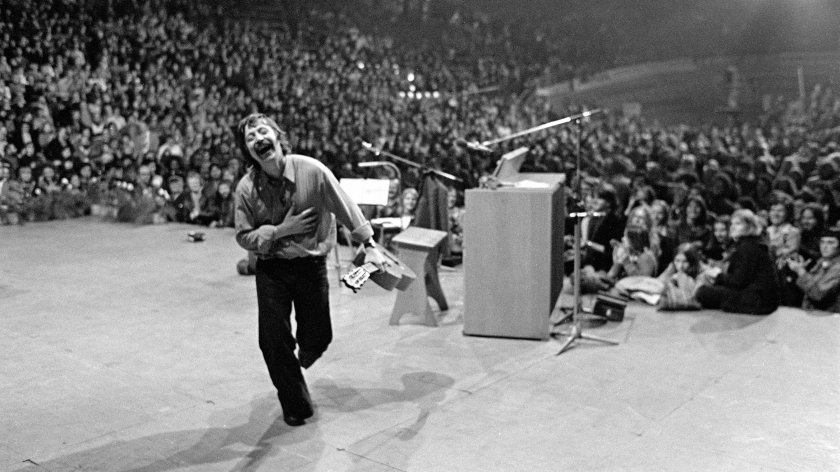 Wolf Biermann mit Mikrofon und Zetteln in der Hand läuft lachend auf einer Bühne vor großem Publikum. Wolf Biermann bei seinem berühmtesten Konzert: In der Kölner Sporthalle im November 1976. Drei Tage spaeter wurde er aus der DDR ausgebuergert.