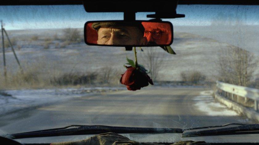 Blick durch die Windschutzscheibe eines Autos auf eine leere Landstraße mit einem hängenden roten Rosenstrauß im Vordergrund und zwei Personen, deren Gesichter im Rückspiegel sichtbar sind