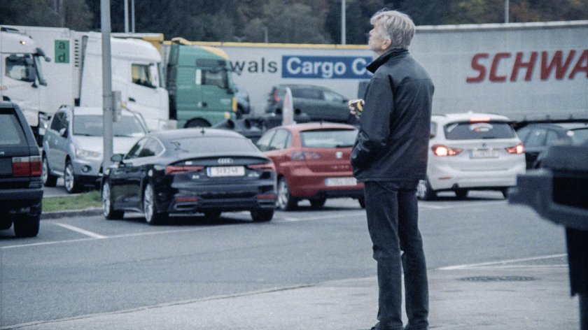 Ein älterer Mann mit grauen Haaren steht auf einem Parkplatz und blickt nach oben, im Hintergrund stehen mehrere Lastwagen und Autos.