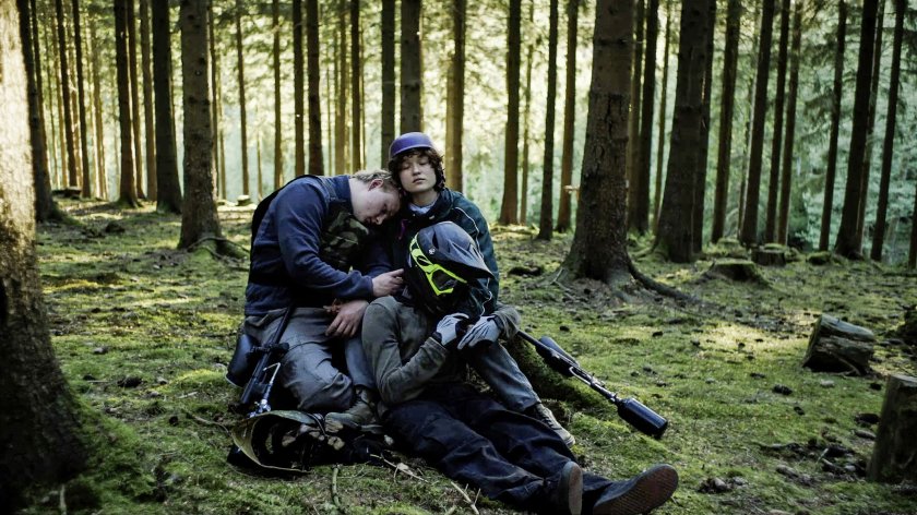 Drei Personen in Outdoor-Kleidung sitzen im Wald auf moosbedecktem Boden, eine Person lehnt den Kopf an die Schulter einer anderen