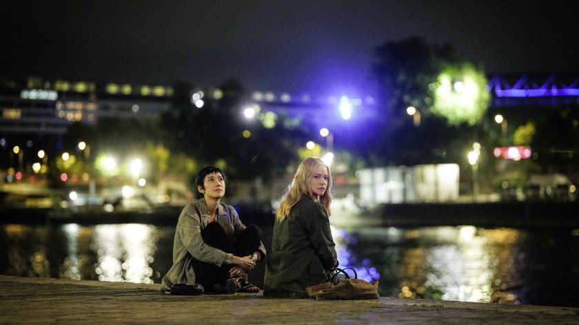 Zwei Frauen sitzen am Boden vor einem Fluss, um sie herum die Lichter der Stadt. Eine der Frauen blickt in die Kamera.