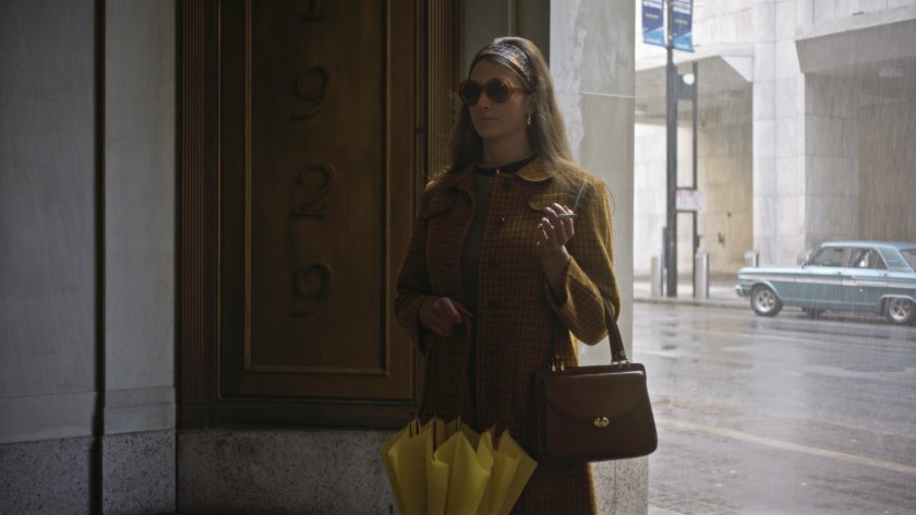 Eine Frau steht in einem Eingangstor, es regnet hinter ihr, sie trägt Sonnenbrille und hat eine Zigarette in der Hand.