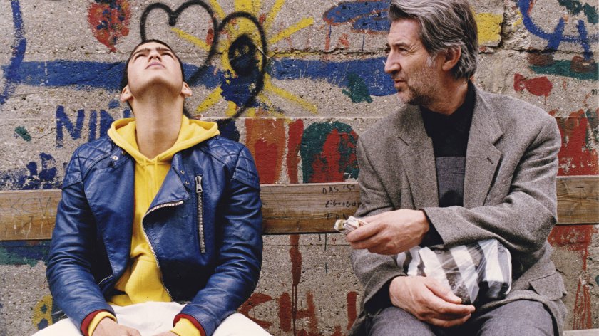 Filmstill JUGOFILM: Zwei Menschen sitzen nebeneinander, hinter ihnen eine mit Graffiti bemalte Wand.