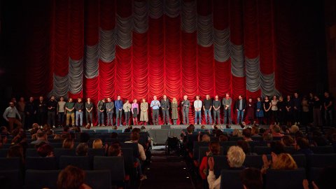 Frontalansicht der Gartenbaukino-Bühne. Der Saal ist voll mit Publikum, auf der Bühne stehen viele Menschen, die dem Publikum zugerichtet sind.