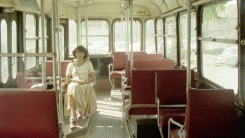 Eine Frau sitzt in einem Bus oder einer Straßenbahn, alle Plätze um sie herum sind leer. Die Sitze sind rot.