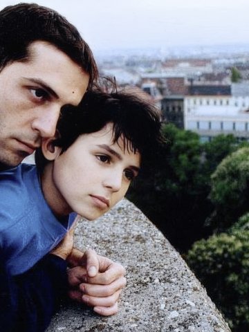 FIlmstill JUGO FILM. Zwei Personen lehnen an einer Mauer und blicken nachdenklich in die Ferne über eine Stadt mit Bäumen und Gebäuden