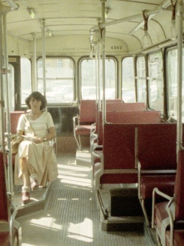 Eine Frau sitzt in einem Bus oder einer Straßenbahn, alle Plätze um sie herum sind leer. Die Sitze sind rot.